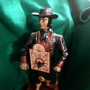 May include: A vintage figurine of a man in a black hat and coat, red vest, and blue pants, holding a clock. The clock face features Roman numerals and a ship illustration. The figurine is on a rectangular base.
