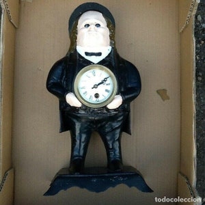 Può includere: Un orologio a forma di statuetta vintage a forma di uomo in abito e cappello neri, che tiene un quadrante rotondo. Il quadrante ha numeri romani e lancette nere. La statuetta è su una base nera.