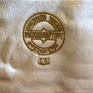Pode incluir: Close-up de um emblema dourado com texto cir&iacute;lico, sobre um tecido texturizado branco sujo. O emblema &eacute; circular com um design central e texto adicional. O tecido tem um brilho sutil.
