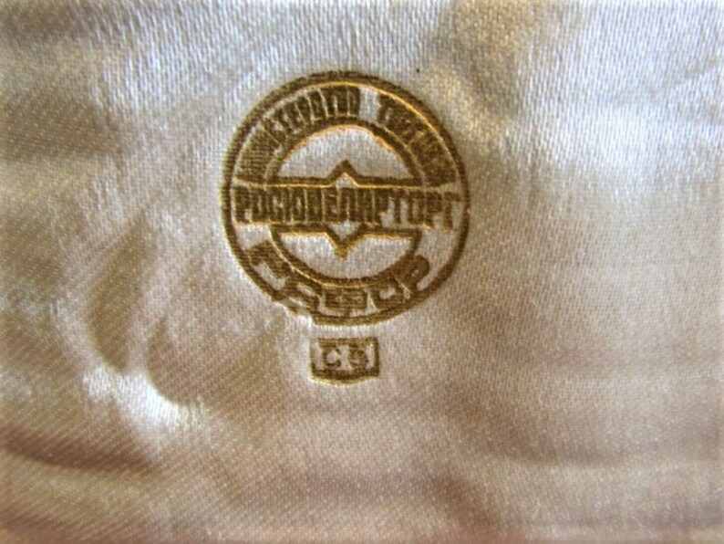 Pode incluir: Close-up de um emblema dourado com texto cir&iacute;lico, sobre um tecido texturizado branco sujo. O emblema &eacute; circular com um design central e texto adicional. O tecido tem um brilho sutil.
