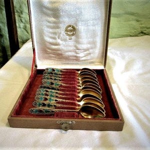 Pode incluir: Um conjunto de colheres douradas com cabos ornamentados em tons de turquesa e preto, exibido numa caixa castanha aberta com interior de veludo vermelho. As colheres est&atilde;o cuidadosamente dispostas. A caixa tem um emblema dourado no interior.