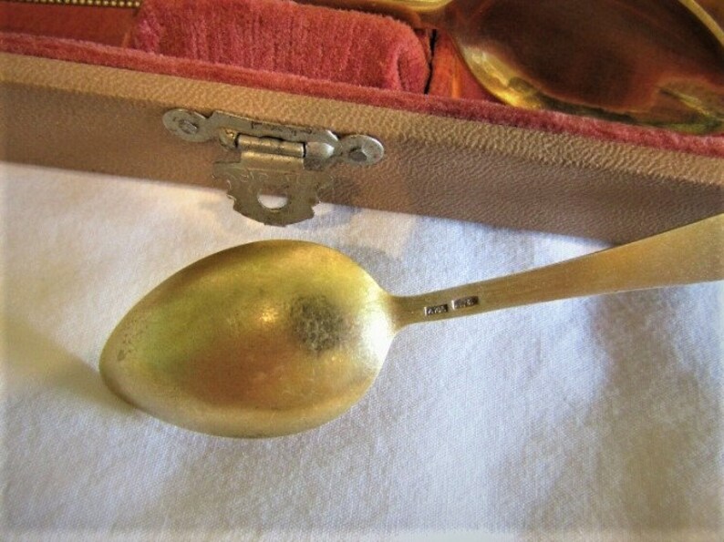 Pode incluir: Close-up de uma colher dourada com uma tigela arredondada e um cabo longo e fino. A colher est&aacute; sobre uma superf&iacute;cie branca, com uma caixa parcialmente vis&iacute;vel forrada com veludo vermelho ao fundo. A al&ccedil;a tem marca&ccedil;&otilde;es.