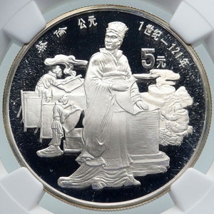 May include: A silver coin featuring a detailed relief of a robed figure with a hat, standing near a table. Other figures are depicted in the background, with Chinese characters and the number "5" visible. The coin is encased in a protective holder.