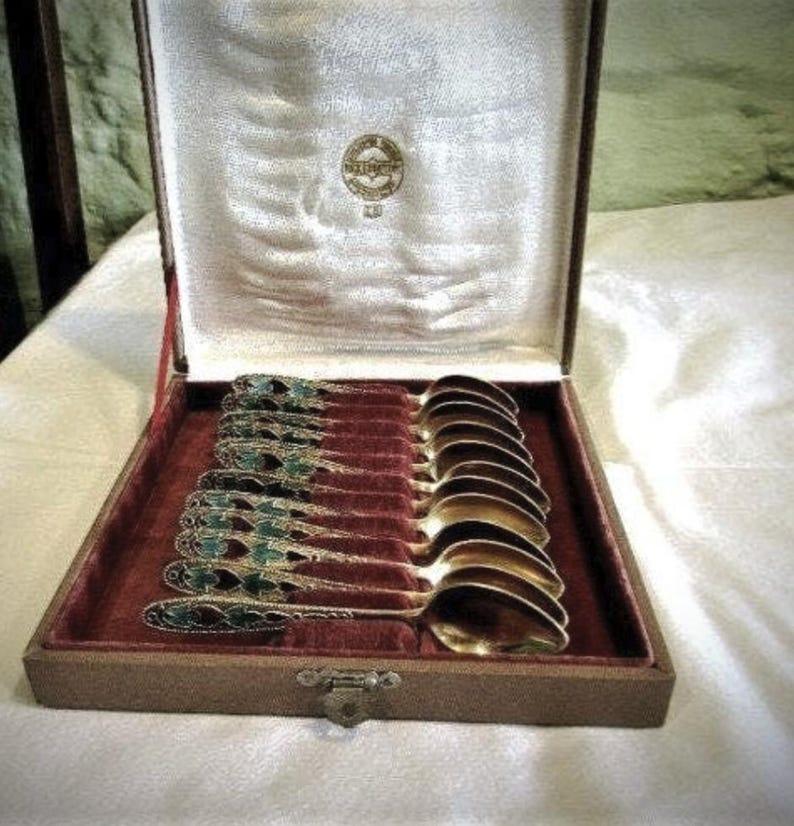 Pode incluir: Um conjunto de colheres antigas numa caixa de madeira. As colheres t&ecirc;m ta&ccedil;as douradas e cabos ornamentados com detalhes em esmalte verde e preto. A caixa tem um forro de veludo vermelho e um fecho de fecho.