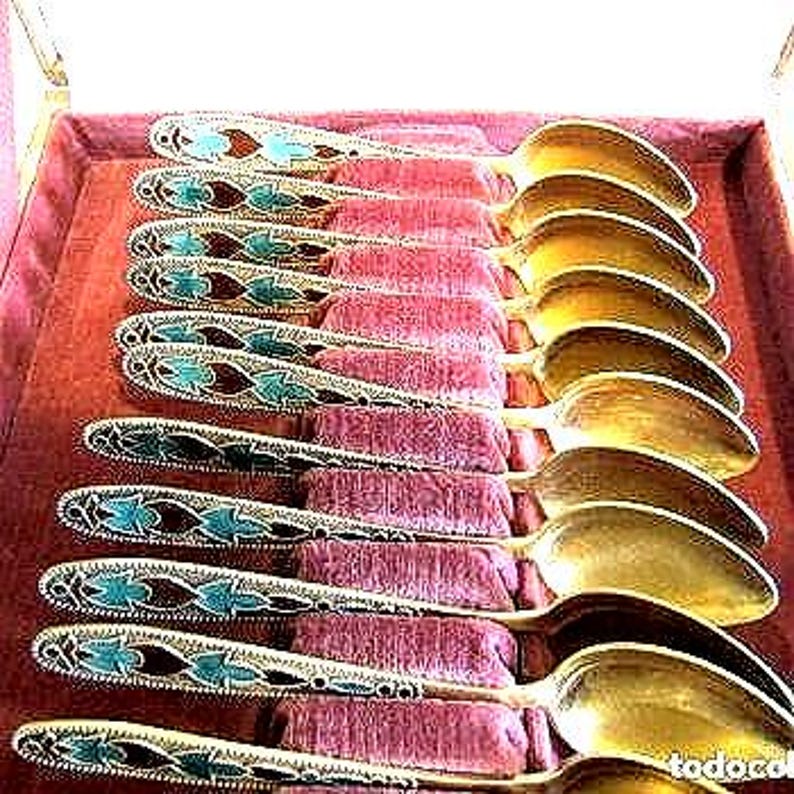 Pode incluir: Um conjunto de colheres antigas douradas com cabos ornamentados com desenhos esmaltados azuis e castanhos. As colheres est&atilde;o dispostas em fila sobre uma superf&iacute;cie de veludo rosa, mostrando o seu artesanato elegante. As colheres medem aproximadamente 18 cm de comprimento.