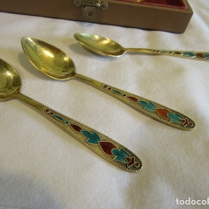 Pode incluir: Tr&ecirc;s colheres douradas com cabos ornamentados. Os cabos s&atilde;o decorados com desenhos em esmalte vermelho, turquesa e dourado. As colheres est&atilde;o dispostas sobre uma superf&iacute;cie branca, com uma caixa de madeira ao fundo.