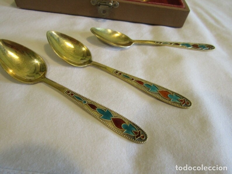 Pode incluir: Tr&ecirc;s colheres douradas com cabos ornamentados. Os cabos s&atilde;o decorados com desenhos em esmalte vermelho, turquesa e dourado. As colheres est&atilde;o dispostas sobre uma superf&iacute;cie branca, com uma caixa de madeira ao fundo.