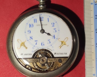 May include: A silver pocket watch with a white face and blue numerals. The watch has gold-colored decorative elements and a visible mechanical movement. The watch is next to a ruler, and the text "8 JOURS" and "8 GUNLUK" are visible.