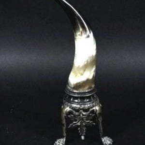 Pode incluir: Um objeto decorativo em forma de chifre com uma base prateada e um chifre curvo e polido. A base apresenta detalhes ornamentados e repousa sobre três pés em forma de garra. O chifre tem um gradiente de cores.