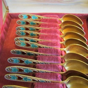 Pode incluir: Um conjunto de colheres douradas com cabos ornamentados, exibido numa caixa forrada a veludo vermelho. Os cabos apresentam um padr&atilde;o decorativo com detalhes turquesa e castanhos, adicionando eleg&acirc;ncia aos talheres.