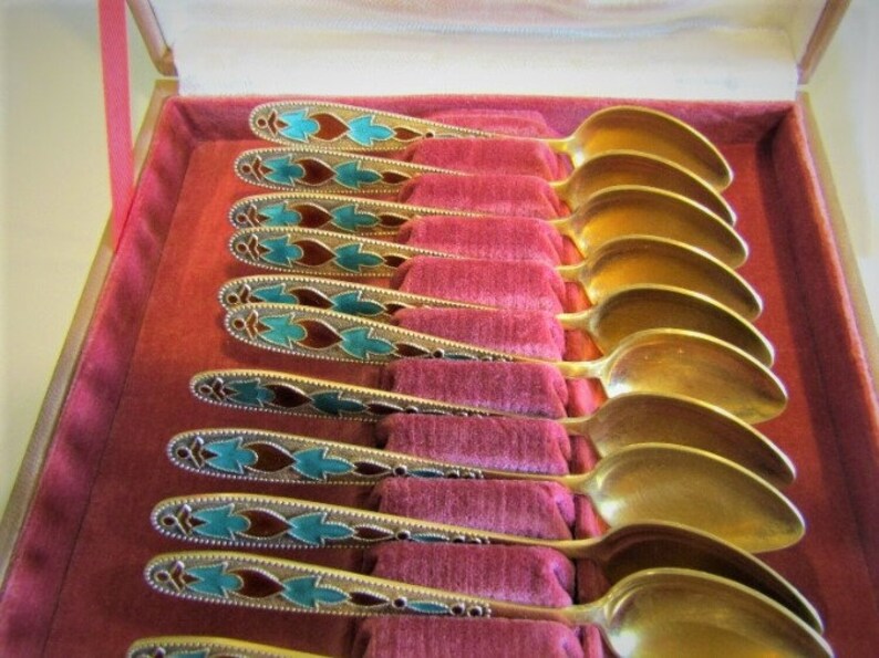 Pode incluir: Um conjunto de colheres douradas com cabos ornamentados, exibido numa caixa forrada a veludo vermelho. Os cabos apresentam um padr&atilde;o decorativo com detalhes turquesa e castanhos, adicionando eleg&acirc;ncia aos talheres.