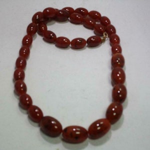 Peut inclure: Un collier composé de perles ovales brun-rouge. Les perles sont enfilées ensemble, formant une boucle complète. Le collier est présenté sur un fond blanc, mettant en valeur la couleur et la forme des perles.