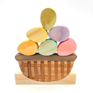May include: A wooden toy basket with a stack of colorful wooden eggs. The basket is brown with a woven pattern and the eggs are yellow, green, purple, pink, and orange.