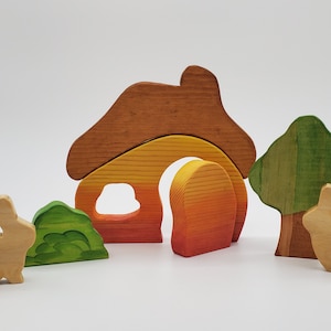 May include: Wooden toy set featuring a house with a brown roof, orange and yellow walls, and a green bush. Two wooden rabbits are included, one standing near the house and one near a green tree.