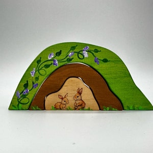 Puede incluir: Un juguete de madera que representa una colina verde con una abertura de cueva marrón. Dos conejos marrones están pintados dentro de la cueva, mirándose el uno al otro. La colina está decorada con enredaderas verdes y flores moradas.