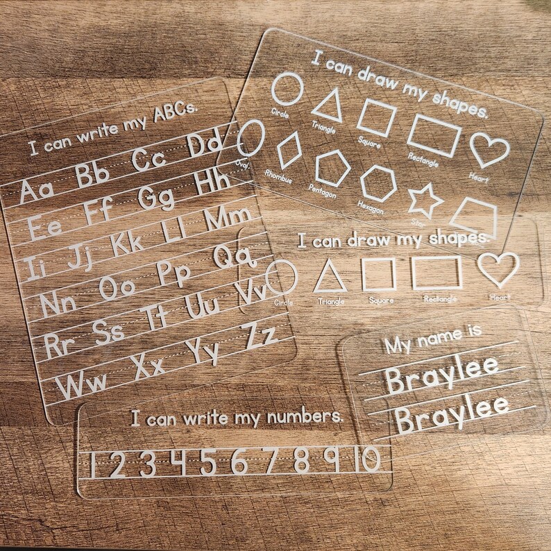 May include: Four clear acrylic dry-erase boards with white lettering. The first board has the alphabet written in rows. The second board has the words "I can draw my shapes." and shows a circle, triangle, square, rectangle, rhombus, pentagon, hexagon, star, and heart. The third board has the words "I can draw my shapes." and shows a circle, triangle, square, rectangle, and heart. The fourth board has the words "My name is" and the name "Braylee" written twice. The last board has the numbers 1 through 10 written in a row.