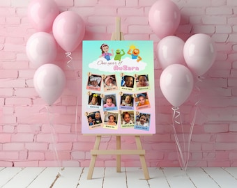 May include: A pink and white photo display board on a wooden easel, featuring a collage of twelve baby photos. The board has the text "One year of AuZara" with cartoon characters and is surrounded by pink balloons.
