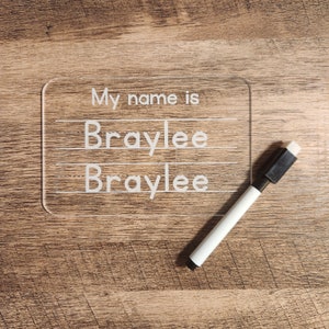 May include: A clear plastic dry-erase board with the text "My name is" and the name "Braylee" written twice. A white dry-erase marker is lying next to the board.