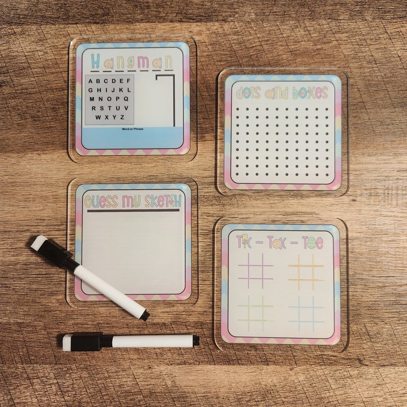 May include: Four colorful dry-erase game boards with pastel pink, blue, and yellow borders. The boards are labeled "Hangman", "Dots and Boxes", "Guess My Sketch", and "Tic-Tac-Toe".
