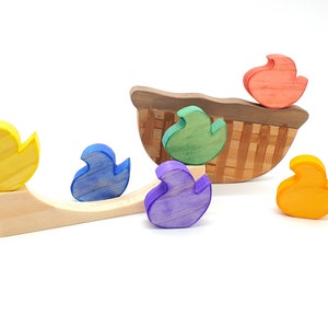 Pode incluir: Um conjunto de brinquedos de madeira com uma cesta e cinco figuras de patos coloridos. Os patos são amarelos, azuis, verdes, roxos e laranja. A cesta é feita de madeira e tem um padrão tecido.