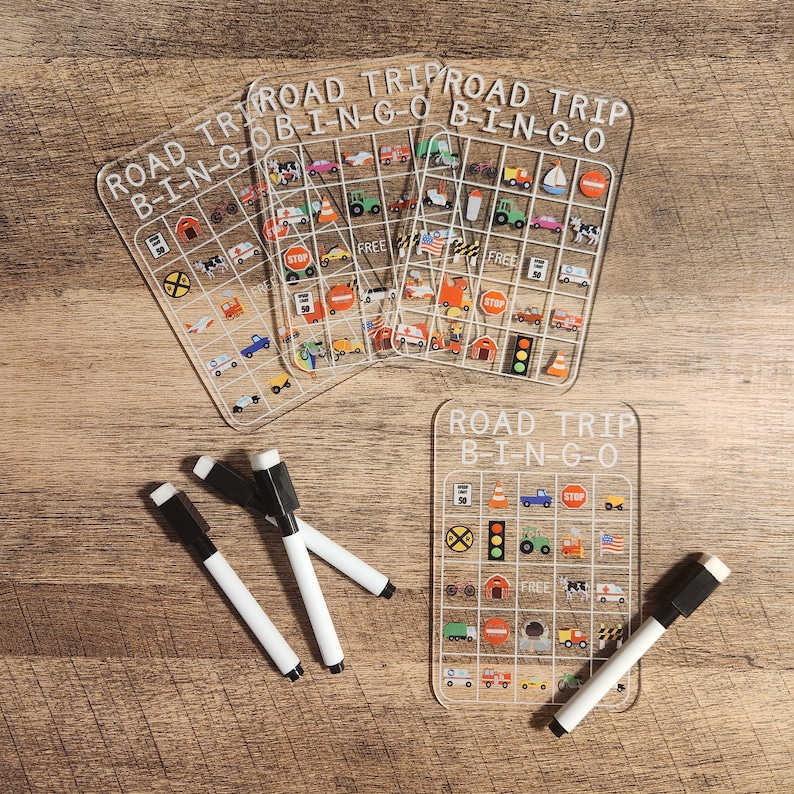 May include: Four clear plastic bingo cards with the text "Road Trip Bingo" and various road trip related images. Three dry erase markers are on the table next to the cards.