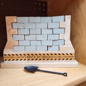 May include: A wooden toy construction set with a brick wall made of gray blocks. The wall has a yellow and black caution tape with the text "BRICK BY BRICK" on it. A small black plastic shovel is included.
