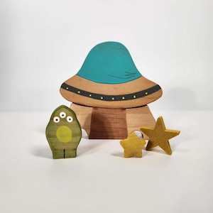 May include: A wooden toy set featuring a blue and brown spaceship, a green alien with three eyes, and two yellow stars. The spaceship has a round top and a flat base. The alien is standing upright with its arms at its sides. The stars are lying on their sides.