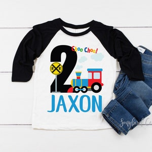 May include: A white and black baseball tee shirt with a blue and red train design. The shirt says "Choo Choo!" and "Jaxon" in blue letters.