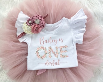 May include: A white onesie with a floral design spelling out "ONE" and the text "Bailey is one-derful". The onesie is paired with a pink tulle tutu and a floral headband.