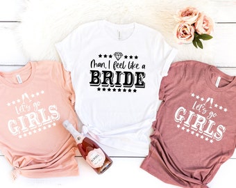 May include: Three t-shirts with text printed on them. One white t-shirt says "Man, I feel like a BRIDE" with a diamond graphic above the text. Two pink t-shirts say "Let's go GIRLS" with a champagne flute graphic above the text.