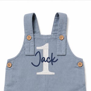 Puede incluir: Un peto de color azul polvoriento con un n&uacute;mero uno grande bordado en blanco y el nombre Jack en letra cursiva azul oscuro. La prenda incluye dos tirantes con botones de madera clara y botones a juego en los laterales.