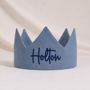 Puede incluir: Una corona de tela en color azul polvoriento con cinco puntas. El nombre Holton est&aacute; bordado en cursiva azul oscuro en la parte central frontal de la corona, que presenta un acabado mate liso y un dise&ntilde;o estructurado y erguido.