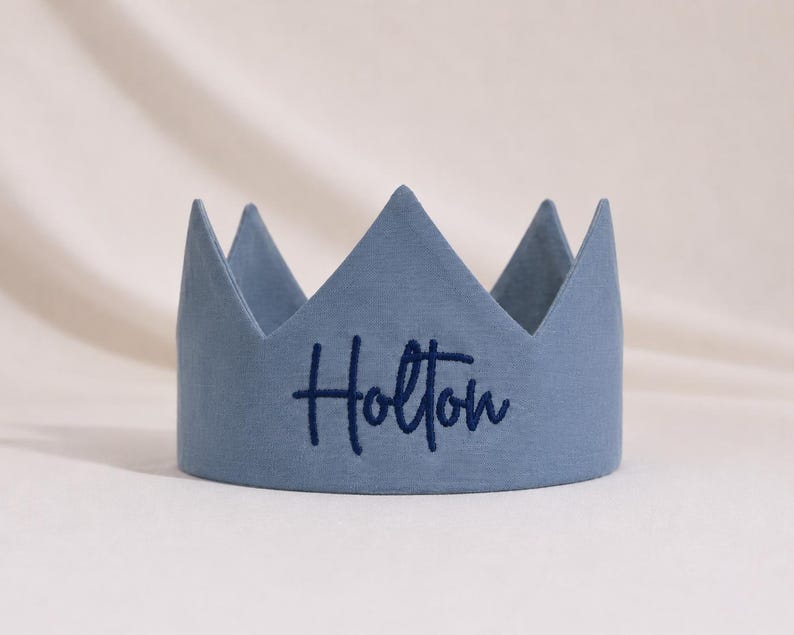 Puede incluir: Una corona de tela en color azul polvoriento con cinco puntas. El nombre Holton est&aacute; bordado en cursiva azul oscuro en la parte central frontal de la corona, que presenta un acabado mate liso y un dise&ntilde;o estructurado y erguido.