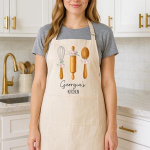 May include: Cream-coloured apron with a pocket, decorated with images of a whisk, rolling pin, and spoon, each with pink floral details. The text "Georgia's KITCHEN" is printed below the illustrations. The apron is worn by a person.