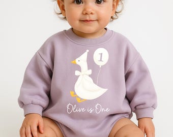 May include: A lavender-colored baby bodysuit with a white goose graphic wearing a party hat and bow tie, holding a balloon with the number 1. The text "Olive is One" is below the goose. The baby is wearing white socks.