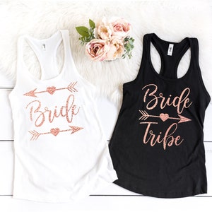 May include: Two tank tops with rose gold glitter text and arrow designs. One white tank top says "Bride" and the other black tank top says "Bride Tribe".
