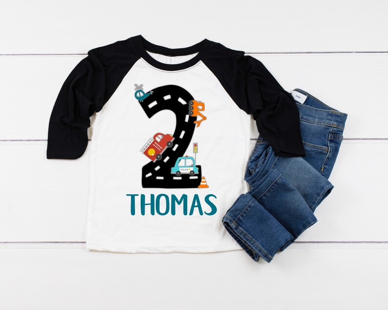 May include: A white and black baseball tee shirt with a number two design featuring a road with cars, a helicopter, and a construction vehicle. The name "THOMAS" is printed in teal below the design.