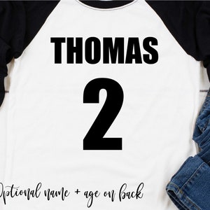 May include: White baseball tee shirt with black sleeves. The front of the shirt has the name "THOMAS" and the number "2" printed in black.