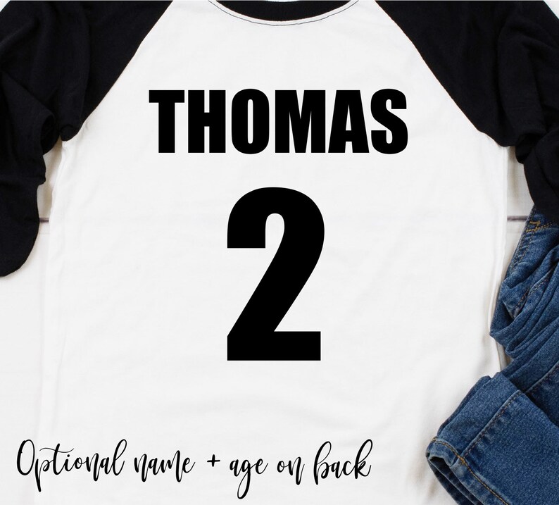 May include: White baseball tee shirt with black sleeves. The front of the shirt has the name "THOMAS" and the number "2" printed in black.