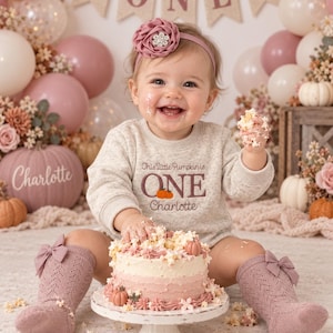 Puede incluir: Una sudadera beige de manga larga con el texto bordado This little Pumpkin is ONE Charlotte y un pequeño gráfico de calabaza naranja. La prenda se lleva con calcetines de punto malva adornados con pequeños lazos y una diadema floral a juego.