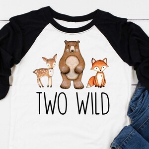 May include: A white and black baseball tee shirt with a brown bear, a deer, and a fox. The shirt says "Two Wild" in black text.