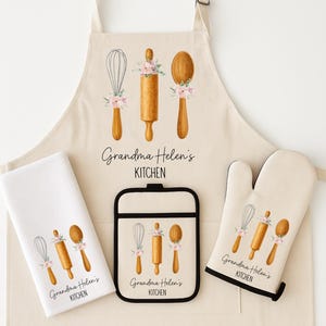 May include: A cream-coloured kitchen set including an apron, oven glove, pot holder, and tea towel. Each item features illustrations of a whisk, rolling pin, and spoon, decorated with pink floral accents. The text "Grandma Helen's KITCHEN" is printed on each item.