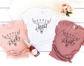 May include: Three t-shirts with a cow skull graphic and the text "Gettin' Hitched" and "Gettin' Rowdy". The shirts are in white, peach, and mauve.