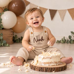 Puede incluir: Un peto de lino beige con botones marrones y el nombre Noah bordado sobre el n&uacute;mero 1. La prenda presenta un escote cuadrado, tirantes anchos y un corte holgado, usado por un ni&ntilde;o peque&ntilde;o sentado junto a un pastel en una tabla.