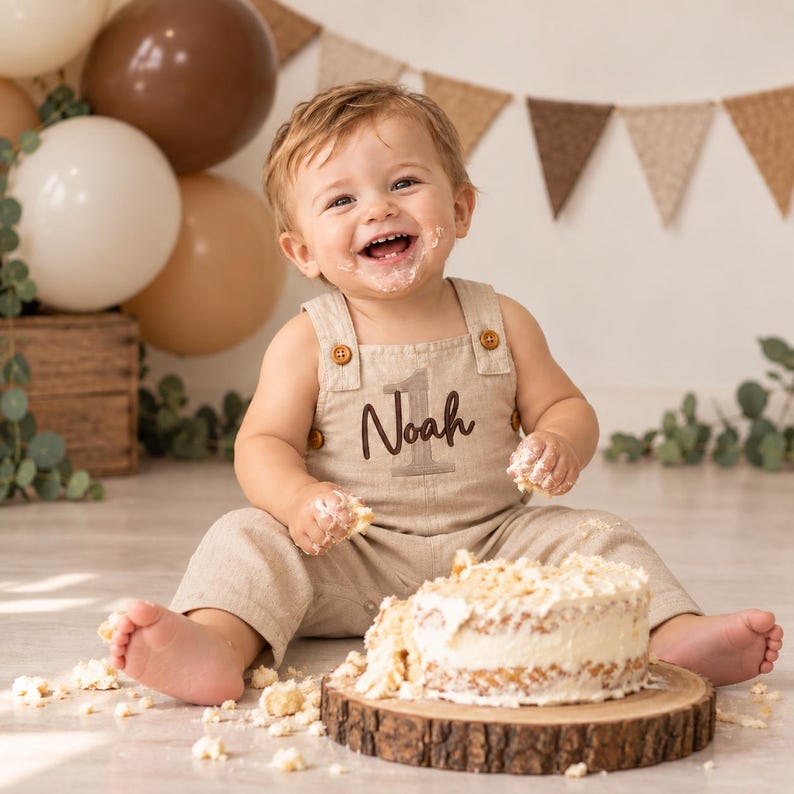 Puede incluir: Un peto de lino beige con botones marrones y el nombre Noah bordado sobre el n&uacute;mero 1. La prenda presenta un escote cuadrado, tirantes anchos y un corte holgado, usado por un ni&ntilde;o peque&ntilde;o sentado junto a un pastel en una tabla.