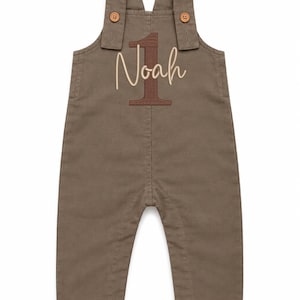 Puede incluir: Un peto de algod&oacute;n color topo con dos botones de madera marr&oacute;n en los tirantes. La zona del pecho muestra un n&uacute;mero uno grande bordado en marr&oacute;n oscuro con el nombre Noah cosido encima en una tipograf&iacute;a cursiva beige claro.