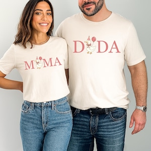 May include: Two cream-colored t-shirts with the words "MOMA" and "DADA" printed in pink. Each shirt features a cartoon chicken wearing a party hat and holding a balloon. The shirts are paired with blue jeans.