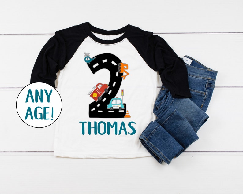 May include: White and black baseball style shirt with a number 2 made of a road with cars and a helicopter on it. The text "THOMAS" is below the number.