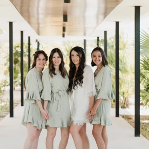 May include: Four women wearing matching sage green robes with white lace trim. The woman in the centre is wearing a white robe with lace trim. They are standing in a hallway with white walls and black pillars.