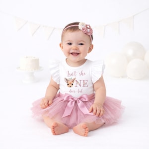 May include: A baby in a white onesie with the text "Isn't she ONE deer-ful" and a deer graphic, paired with a pink tutu skirt and a matching bow. The baby wears a pink floral headband. White balloons and a small cake are in the background.